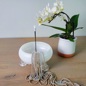 May include: A white ceramic bowl with a metal rod and a string of beige and white beads. A white ceramic pot with an orange rim holds a white orchid plant with green leaves.