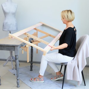 Puede incluir: Una mujer se sienta en una mesa usando un soporte de aro de bordado de madera. El soporte está sujeto a la mesa con una abrazadera. El aro está inclinado en un ángulo, lo que permite a la mujer trabajar cómodamente en su proyecto de bordado.