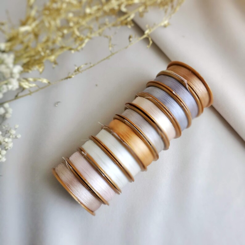 May include: A stack of spools of thread in various shades of beige, white, and grey. The spools are arranged in a graduated colour pattern, with the darkest shade at the bottom and the lightest shade at the top.