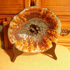 May include: A large, ceramic bowl with a brown, orange, and blue glaze. The bowl has a unique, textured surface and is displayed on a black stand.