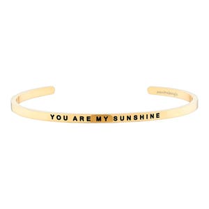 May include: Gold-colored cuff bracelet with the inscription "YOU ARE MY SUNSHINE" in black lettering. The bracelet has a simple, open design and is likely made of metal. The brand name "mantrabangle" is also visible.