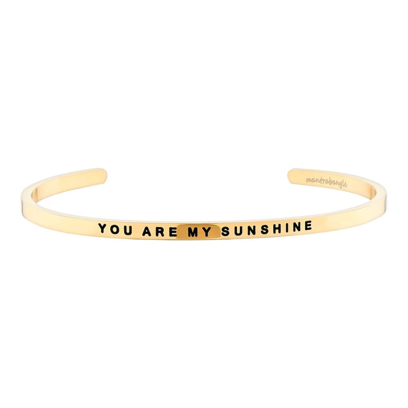 May include: Gold-colored cuff bracelet with the inscription "YOU ARE MY SUNSHINE" in black lettering. The bracelet has a simple, open design and is likely made of metal. The brand name "mantrabangle" is also visible.