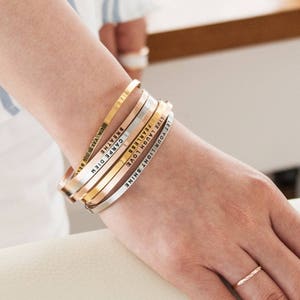 May include: A collection of six metal bangles in gold, rose gold, and silver tones. Each bangle features an engraved inspirational word or phrase, including "LET YOUR LIGHT SHINE" and "CARPE DIEM". A simple gold ring is also visible.
