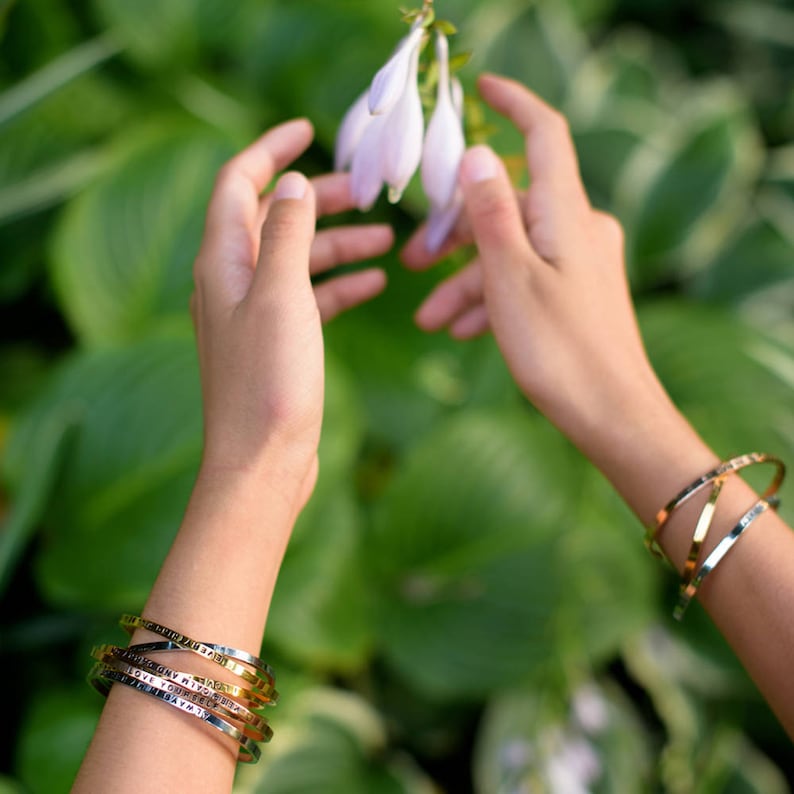 May include: A collection of gold and silver metal bracelets with engraved text. The bracelets are stacked on a wrist, with a second wrist wearing a gold bracelet. The background features green foliage and a person's hands reaching for white flowers.