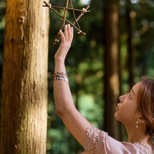 May include: A wooden star made of twigs is held up by a person's hand. The person is wearing multiple bracelets on their arm. The background is a forest with tall trees and green foliage. The person is wearing a pink lace top.