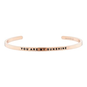 May include: Rose gold cuff bracelet with the words "YOU ARE MY SUNSHINE" engraved in black. The bracelet is a simple, open design with rounded edges. The brand name "mantrabangle" is also visible.