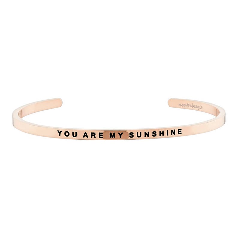 May include: Rose gold cuff bracelet with the words "YOU ARE MY SUNSHINE" engraved in black. The bracelet is a simple, open design with rounded edges. The brand name "mantrabangle" is also visible.