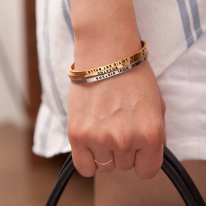 May include: A gold, silver, and gold-tone bangle bracelet set. The gold bracelet has the words "LA VIE EST BELLE" engraved on it. The silver bracelet has the words "BREATHE" engraved on it. The gold bracelet has the words "SOUL SISTERS" engraved on it.