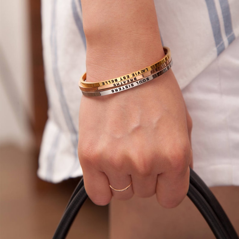 May include: A gold, silver, and gold-tone bangle bracelet set. The gold bracelet has the words "LA VIE EST BELLE" engraved on it. The silver bracelet has the words "BREATHE" engraved on it. The gold bracelet has the words "SOUL SISTERS" engraved on it.