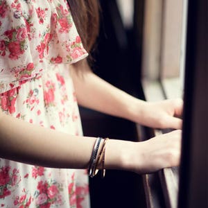 May include: A set of three thin bangles in silver and gold tones. The bangles are worn on a person's arm, with a floral print dress in the background. The dress has a white base with red and pink roses.