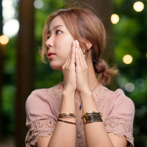 May include: A woman wearing a pink lace-sleeved top and multiple gold and black bracelets. The bracelets are stacked and feature black accents. The woman's hands are clasped together in front of her, with a blurred green background.