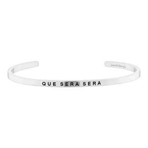 May include: A silver-colored cuff bracelet with the phrase "QUE SERA SERA" engraved in black lettering. The bracelet has a minimalist design and the brand name "mantrabangle" is also visible.