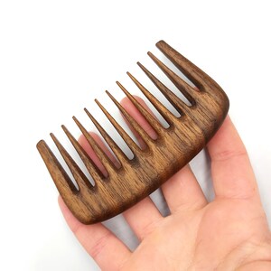 Puede incluir: Un peine de madera marr&oacute;n oscuro con dientes anchos. El peine se sostiene en una mano.
