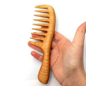 Pode incluir: Um pente de madeira com acabamento natural e cabo curvo. O pente tem dentes largos e &eacute; projetado para desembara&ccedil;ar o cabelo.