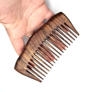 K&ouml;nnte beinhalten: Ein dunkelbrauner Holzkamm mit einem breiten, abgerundeten Griff und einer Reihe feiner Z&auml;hne. Der Kamm wird in einer Hand gehalten, wodurch seine Gr&ouml;&szlig;e und Form deutlich werden.