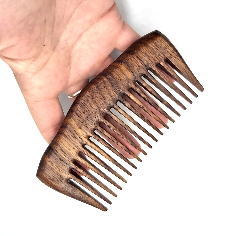 K&ouml;nnte beinhalten: Ein dunkelbrauner Holzkamm mit einem breiten, abgerundeten Griff und einer Reihe feiner Z&auml;hne. Der Kamm wird in einer Hand gehalten, wodurch seine Gr&ouml;&szlig;e und Form deutlich werden.