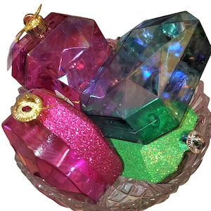 May include: A collection of jewel-shaped Christmas ornaments in a clear glass bowl. The ornaments are various colors, including pink, green, and blue. Some have glittered surfaces, and all have gold-colored caps.