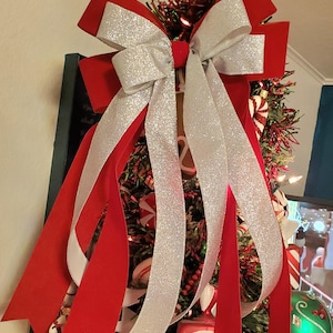 May include: A festive Christmas tree topper featuring a large red velvet bow with silver glitter ribbon accents. The bow is adorned with long, flowing ribbons and is set against a backdrop of a decorated Christmas tree with candy cane ornaments.