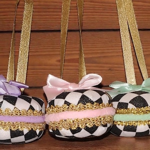 May include: Three decorative ornaments resembling macarons. Each has a black and white checkered pattern, a gold trim, and a ribbon bow. The bows are lavender, pink, and mint green. Gold ribbon loops are attached for hanging.