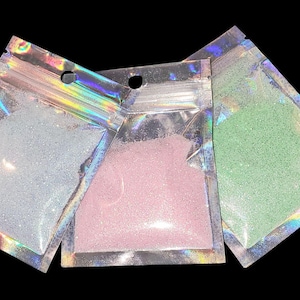 May include: Three small, clear, holographic zip-lock bags filled with glitter. The glitter colors are light blue, pink, and green. The bags are arranged in a fan shape against a black background.