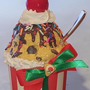 May include: A decorative cupcake ornament featuring a yellow base with chocolate chips, topped with white frosting, chocolate drizzle, colorful sprinkles, and a red cherry. It is wrapped in a red and white striped paper liner with a green ribbon bow.