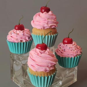 May include: Four cupcakes in teal paper liners, each topped with a swirl of pink frosting, colorful sprinkles, and a single red cherry. The cupcakes are arranged on a clear, tiered acrylic display stand against a neutral background.
