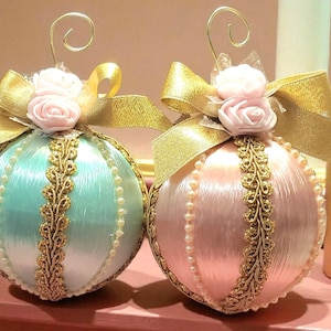 May include: Two decorative Christmas ornaments. One is light blue, the other is pink. Both are round with gold ribbon, pearl accents, and gold filigree. Each has a small cluster of pink roses and a gold wire hanger.