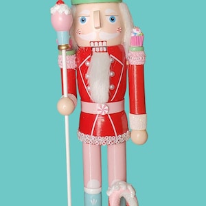 May include: A whimsical Christmas nutcracker figurine with a cupcake-shaped hat, red jacket, and pink legs. The figurine holds a staff with a cupcake topper and stands on a base with candy cane and peppermint accents.