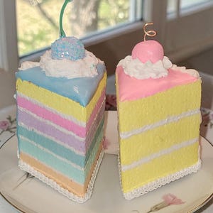 May include: Two decorative cake slices, one pastel rainbow and the other yellow with pink frosting. Each slice is topped with a decorative cherry and sits on a floral patterned plate. The cakes have a textured appearance.