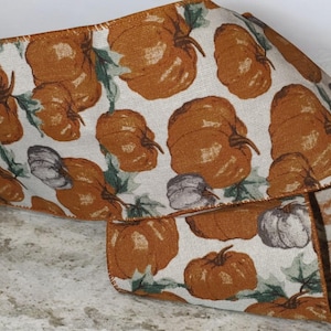 May include: Decorative ribbon featuring a repeating pattern of orange pumpkins and gray gourds on a cream background. Green leaves accent the pumpkins. The ribbon is folded, showing the pattern on multiple sides.