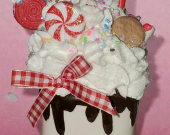 May include: A whimsical, marshmallow-shaped container with a smiling face, topped with white whipped cream, candies, and a red and white checkered bow. The container is decorated with chocolate drips.