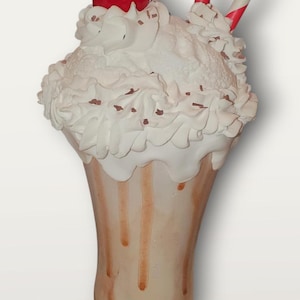 May include: A decorative milkshake with a red cherry on top, whipped cream, and two red and white striped straws. The milkshake glass has brown drips and a decorative base with a checkered bow.