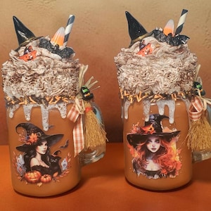 May include: Two glass mugs filled with orange liquid, topped with faux whipped cream, black witch hats, candy corn, and striped straws. Each mug features a witch illustration, orange sprinkles, and a decorative raffia broom tied with a plaid ribbon.