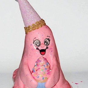 May include: A pink, cartoon-like figure with a party hat and a cupcake. The figure has a smiling face and is holding a cupcake with colorful sprinkles. The party hat is light pink with glitter and a gold trim.