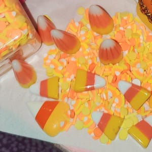 May include: A collection of candy corn-themed craft supplies. The image features candy corn-shaped embellishments in orange, yellow, and white, along with a container of confetti in the same colors. The items are scattered on a white surface.
