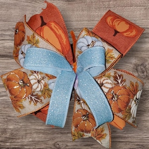 May include: A decorative fall-themed bow featuring orange and white pumpkins, fall leaves, and a light blue glitter ribbon. The bow is made of various ribbons and is suitable for autumn decorations.