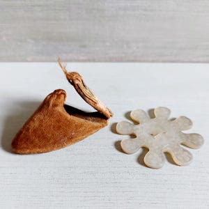 May include: A brown velvet slipper with a dried flower and a white, flower-shaped button. The slipper is angled, showing its interior. The button has a small hole in the center.