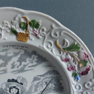 May include: White ceramic plate with a detailed black and white pastoral scene. The plate's rim features embossed floral and vine designs with touches of green, purple, and gold. The word "Vergn&uuml;gen" is visible on the plate.