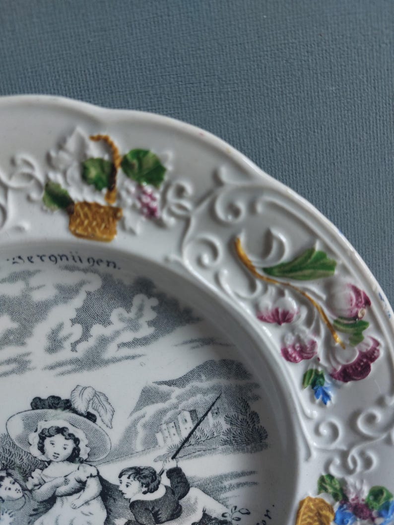 May include: White ceramic plate with a detailed black and white pastoral scene. The plate's rim features embossed floral and vine designs with touches of green, purple, and gold. The word "Vergn&uuml;gen" is visible on the plate.