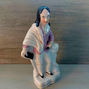 Könnte beinhalten: Vintage Porzellanfigur eines Mannes in Kleidung des 18. Jahrhunderts. Die Figur trägt ein weißes Hemd, eine lila Jacke und weiße Hosen. Ein weißer Schal ist über den Schultern drapiert. Die Figur steht auf einem weißen Sockel.