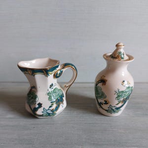 Op de afbeelding: Een witte keramische creamer en pot met deksel set. De creamer heeft een teal handvat en rand met gouden accenten. Beide stukken zijn versierd met teal bloempatronen en gouden details. De set is waarschijnlijk voor het serveren van thee of koffie.