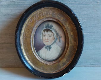 Puede incluir: Miniatura de retrato ovalada de una mujer con un gorro y vestido blancos, con una cinta azul. La pintura está enmarcada en un marco ovalado negro y dorado con un lazo para colgar.