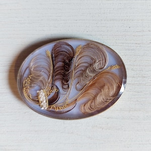 May include: An oval brooch featuring a light blue translucent background encased in a silver-toned metal frame. The interior contains intricate arrangements of brown and blonde hair shaped into feather-like plumes, accented with gold wire and small white beads.