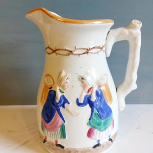 May include: Cream ceramic pitcher with a decorative handle and spout. The pitcher has a brown barbed wire design around the neck and two raised relief figures in blue, green, and pink clothing. The rim is trimmed in gold.