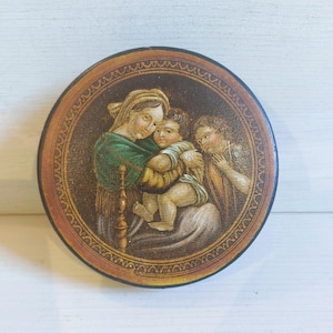 May include: Circular, antique painted wooden box with a religious scene. The artwork shows a woman in a green shawl holding a child, with another figure to the right. The box has a decorative border and a dark brown finish.
