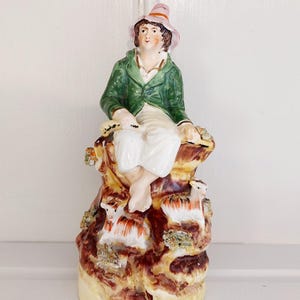 May include: A vintage porcelain figurine depicting a seated figure in a green jacket and pink hat atop a rocky base. The base features sheep and floral accents, with a cream-colored bottom. The figurine is approximately 8 inches tall.