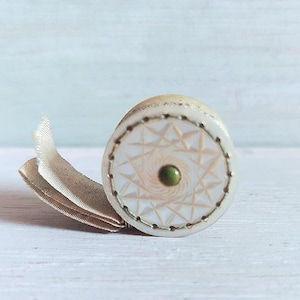 May include: A small, round, off-white decorative box with a geometric starburst pattern on top. The box has a green center and is trimmed with a dark border. A beige ribbon is attached to the side.