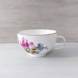 Könnte beinhalten: Weiße Teetasse mit einem Blumendesign aus rosa, gelben und blauen Blumen. Die Tasse hat einen braunen Rand und einen gebogenen Henkel. Die Teetasse steht vor einem hellen Hintergrund.