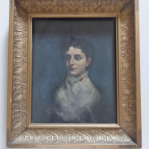 May include: An oil painting portrait of a person with dark hair, wearing a white lace collar with a small red accent, set against a dark blue background. The artwork is enclosed in a rectangular, ornate gold-toned frame with carved floral motifs.