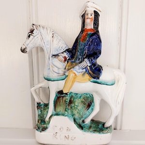 Könnte beinhalten: Keramikfigur eines Mannes auf einem Pferd, gekleidet in Dreispitz und blauem Mantel. Die Figur ist in Weiß, Blau, Gelb und Rot bemalt. Auf dem Sockel der Figur steht der Schriftzug "TOM KING".